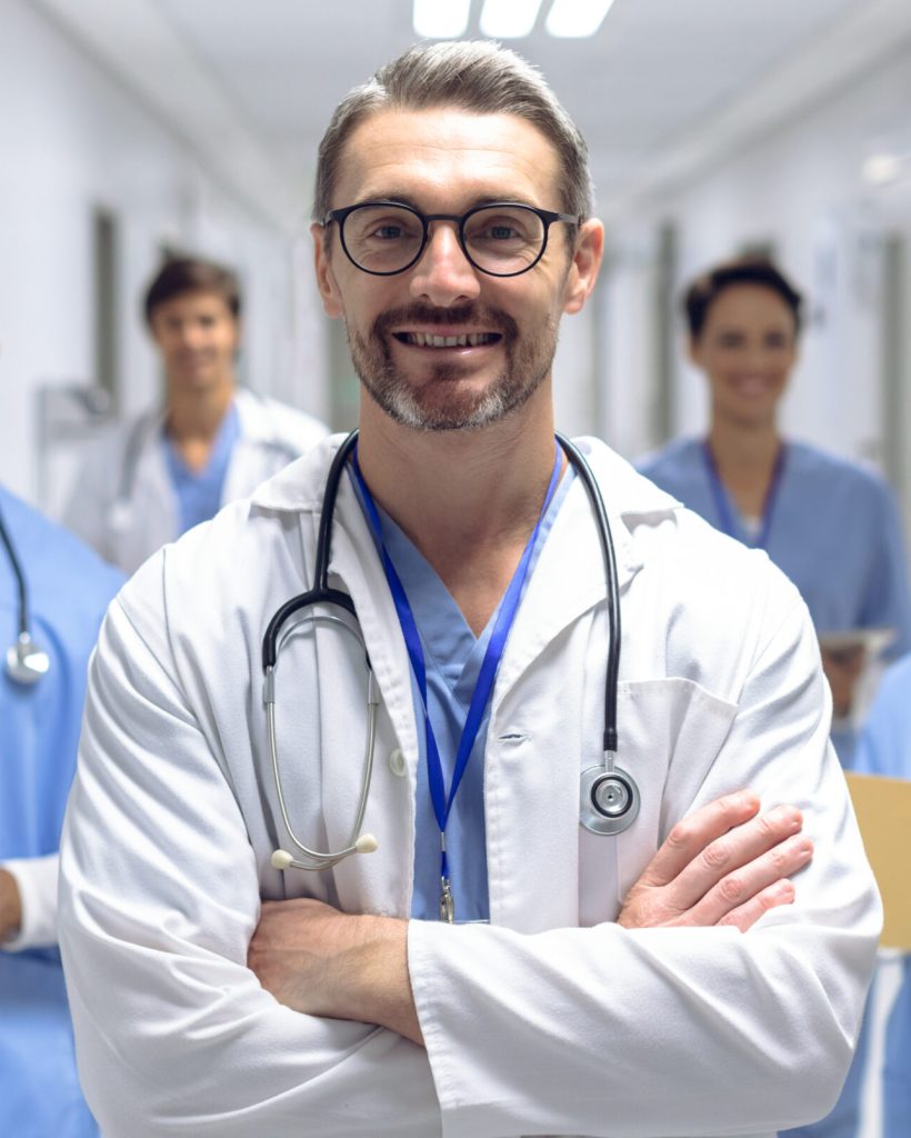 Front view of diverse medical team of doctors looking at camera while holding clipboard and medical files in corridor at hospital. Caucasian male surgeon has his arms crossed.